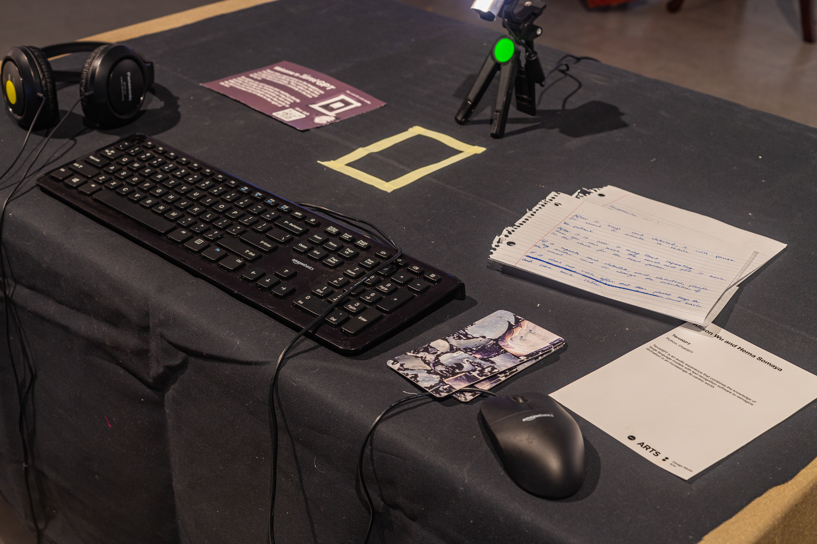 Detailed close up of a table with black table cloth, headphones, a keyboard, a mouse, playing cards and an instruction manual on top. There is a webcam connected to a 6 inch tripod pointing towards the cards.